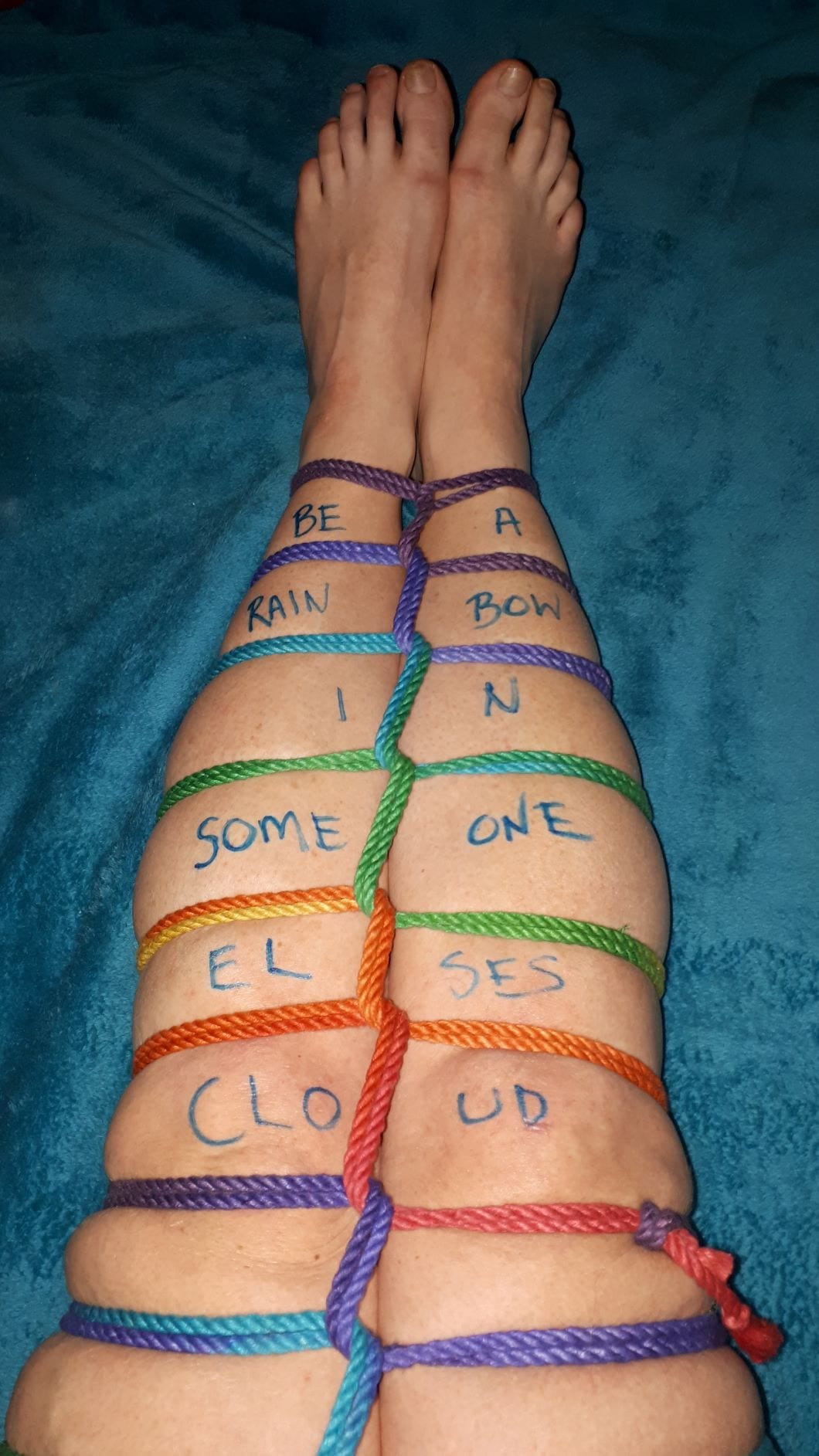 Bare legs bound in rainbow rope with the quote "Be a rainbow in someone else's cloud" written on the shins