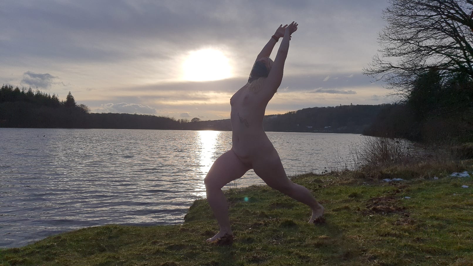 Naked lady doing outdoor yoga: Lakeside warrior pose