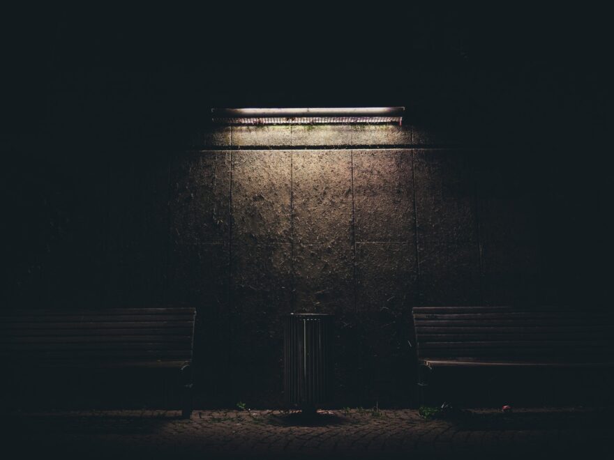 A dark brick wall, a bin in the middle with a bench either side, illuminated from above by a single lightsource.