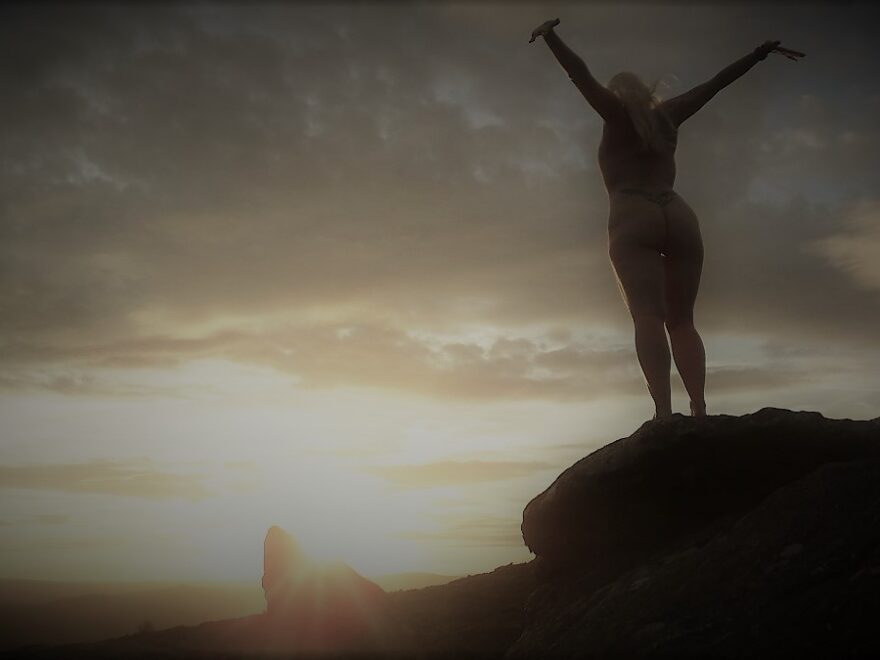 Changing nature of explorations shows naked lady standing on a rock facing away from the camera watching the sunset and reaching up and out with her arms.