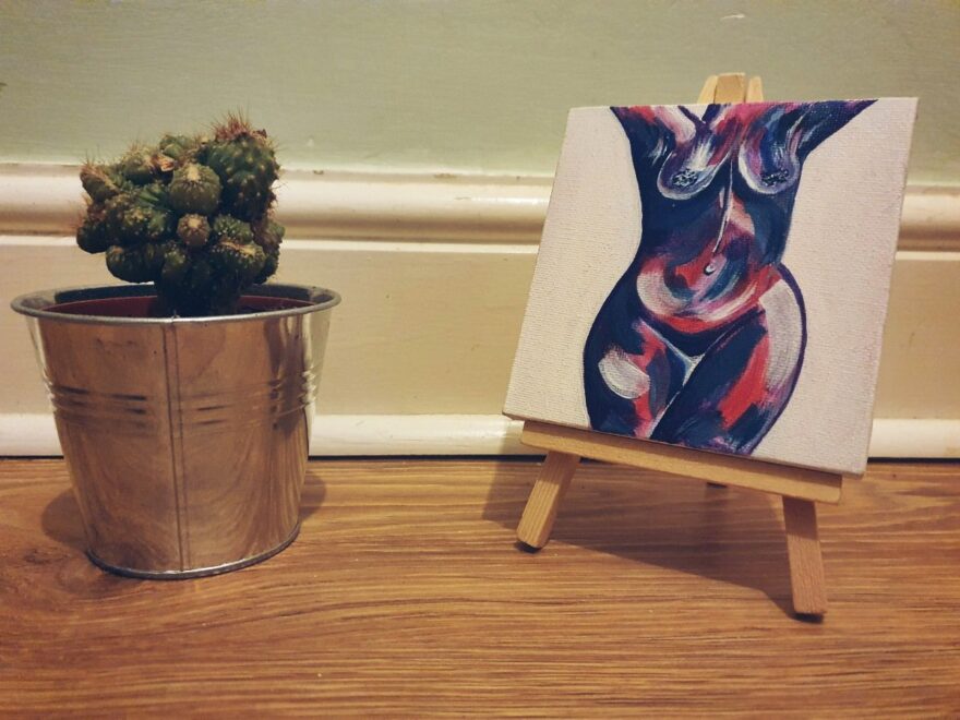 Body confidence mini nudie painting (in dark purple and pink) on a mini easel in front of a skirting board, beside a mini cactus.