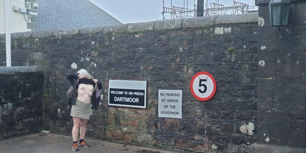 Topless adventure on the outside of the walls of Dartmoor Prison. Blonde lady exposing her breasts beside the prison signage.