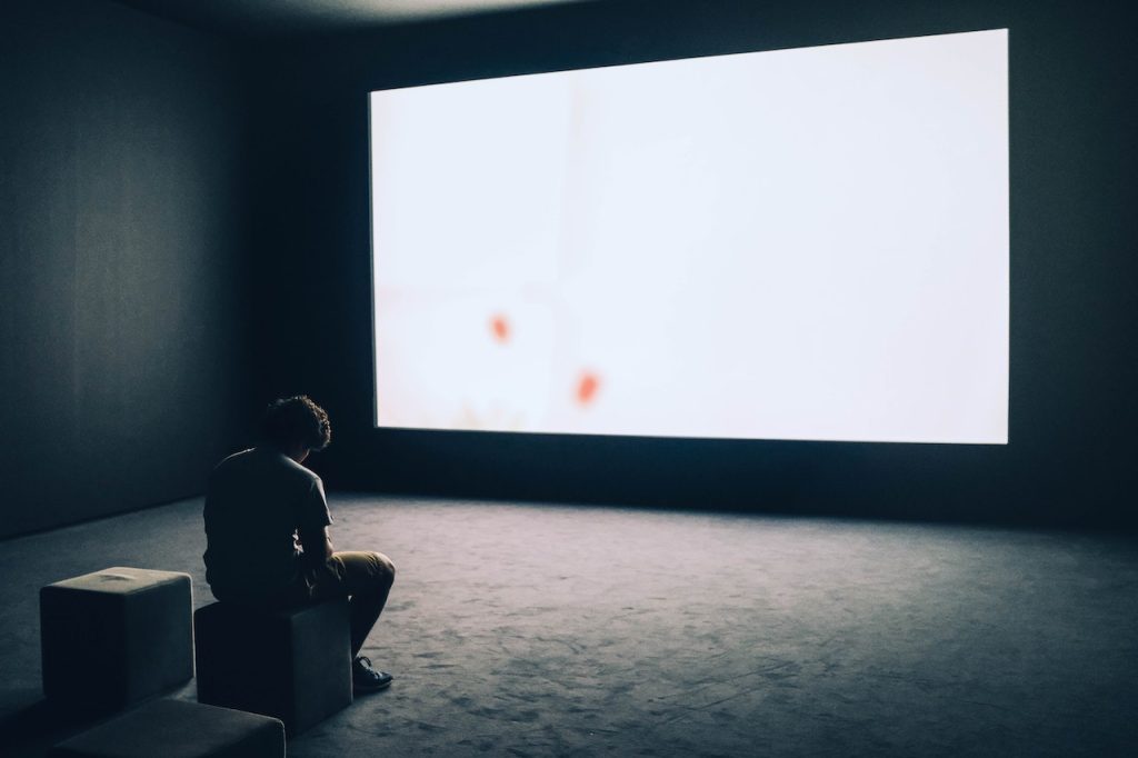 "I Think Part Of Me Will Always Be Waiting For You" header image shows a Person sitting in front of a big white screen in an otherwise empty room.