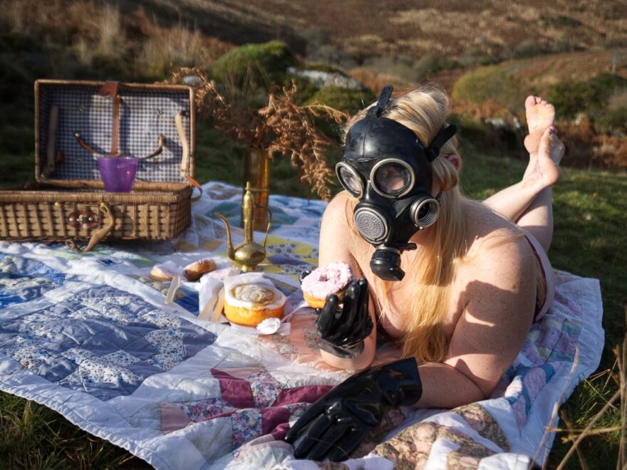 Barefoot, wearing gas mask, corset and latex gloves, is attempting to eat a cake.