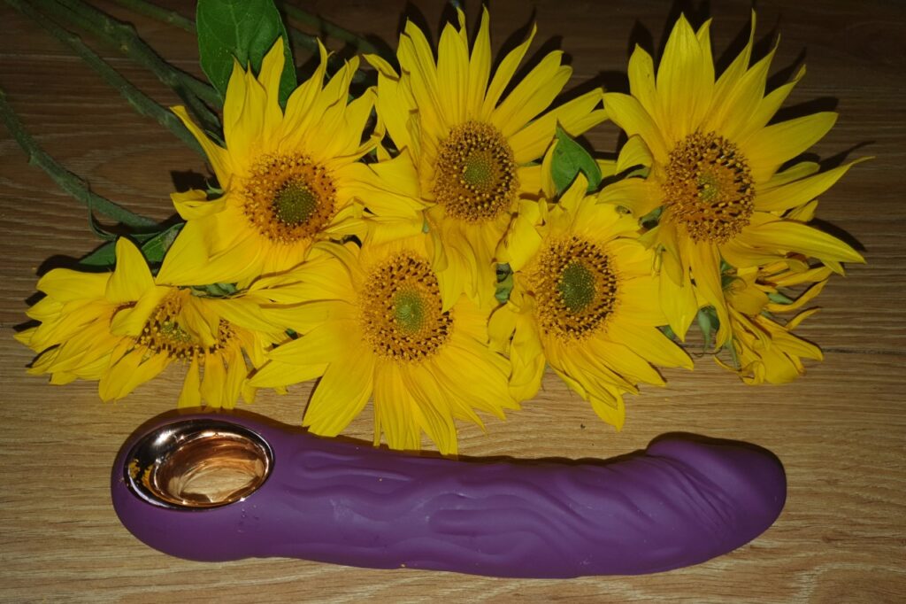 The purple, realistic vibrator with finger loop, laying in front of some sunflowers.